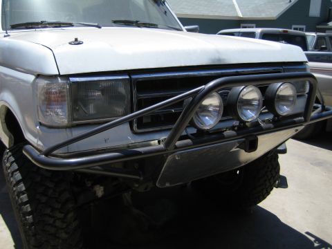1990 Ford Bronco - Front three-quarter view of a white Ford truck with a black tubular prerunner bumper, three round driving lights, large mud tire and raised…