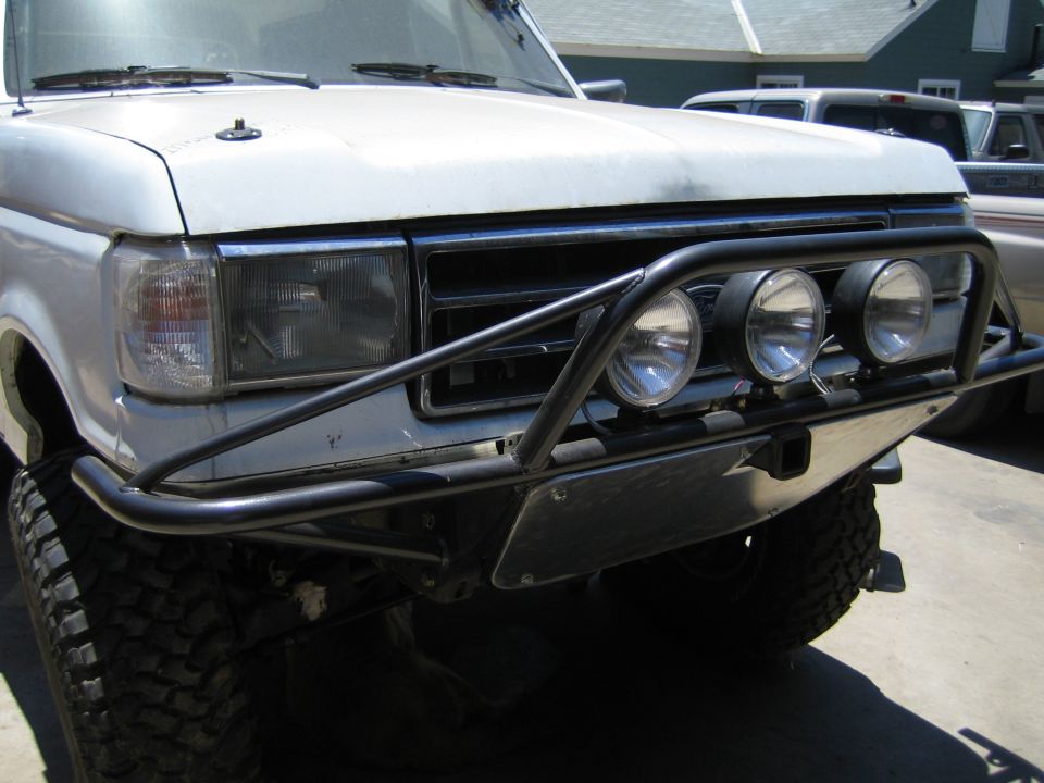 Front three-quarter of a white Ford truck with black tube bumper and three round driving lights