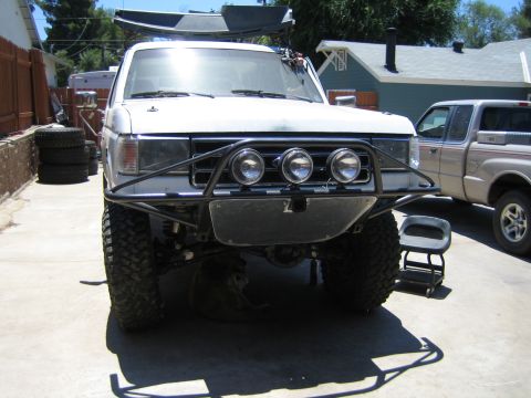 1990 Ford Bronco - Front view of a lifted white Ford truck with a black tubular bumper, three round driving lights and a roof-mounted light bar, parked in a d…