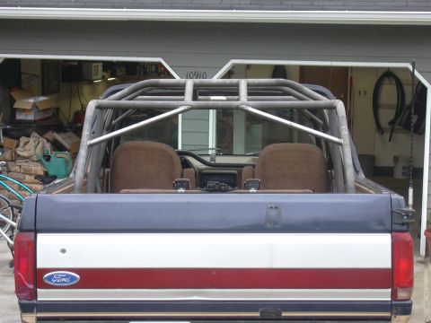1991 Ford Bronco - Rear view of a two-tone blue-over-red-and-white Ford truck with a custom tubular roll cage/roll bar visible over the tan bucket seats and o…