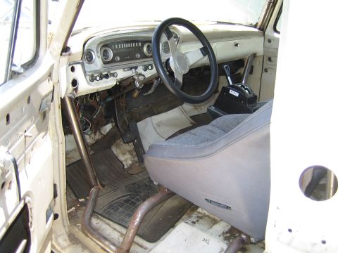1990 Ford Bronco - Worn interior of a vintage pickup truck with chrome dash, round gauges, floor shifter and a gray bench seat over a rusty floor.