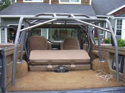 1991 Ford Bronco - Interior rear view of a Ford Bronco looking forward from the cargo area, showing a custom multi-point roll cage in gray/primer finish, tan/…