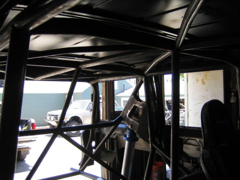 1990 Ford Bronco - Inside a caged vehicle looking out through the windows toward parked pickups, showing an extensive black tube roll cage.