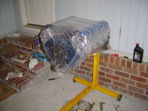 1986 Ford Bronco - An engine block, wrapped in plastic, sits on a yellow engine stand outside a building.