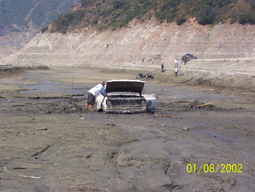 White truck stuck in deep mud with hood up, man working on it, dried lakebed and hills behind
