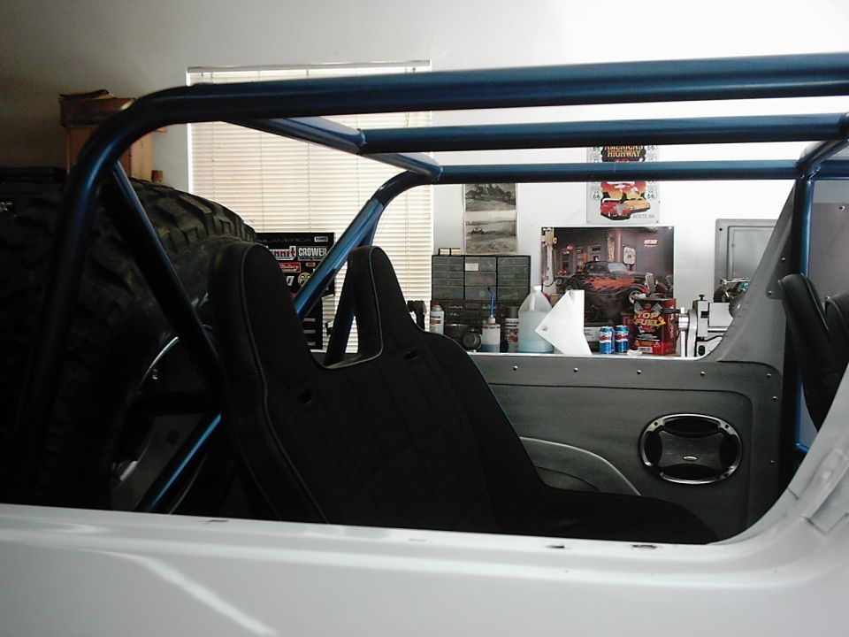 Interior of a white vehicle with a blue roll cage, black bucket seats, and a spare tire visible