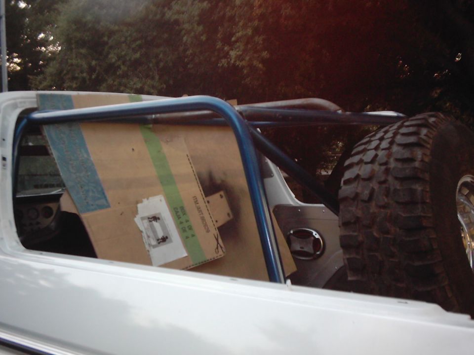 SUV interior with roll cage tubing and cardboard covering part of the window. Spare tire is visible