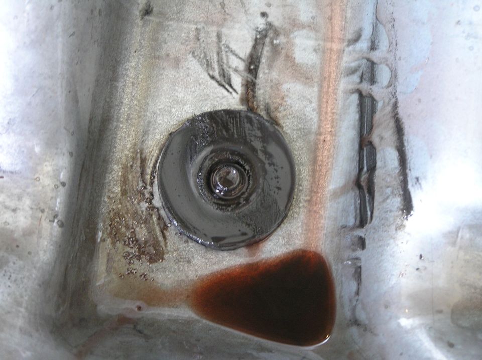 Close-up of metal drain pan with central plug and dark used fluid and rust residue