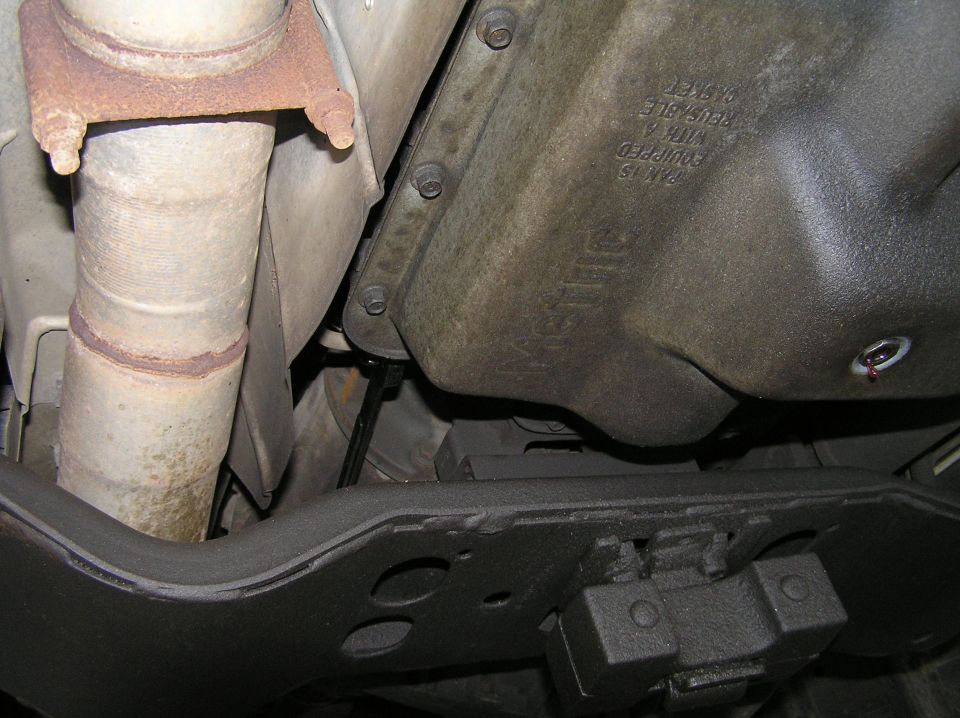 Underside view of black transmission pan with embossed text beside a rusty exhaust pipe and black crossmember.