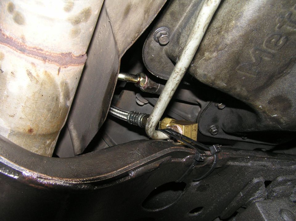 Underside of vehicle showing threaded stud, white line, brass fitting and zip ties near rusty components