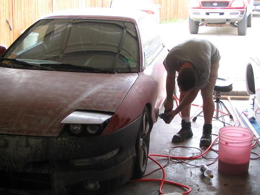 Red 1994 Ford Probe with pop-up headlights and primer, being sanded by a person in a garage