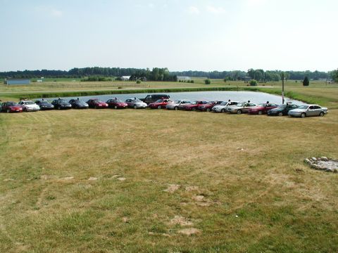 1992 Ford Taurus - A collection of vehicles are lined up in a grassy field next to a body of water under a clear sky.