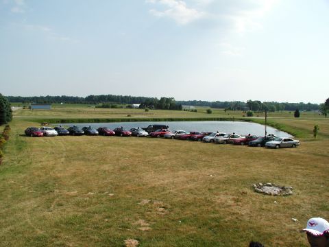 1992 Ford Taurus - A collection of vehicles are parked in a grassy field next to a pond under a partly cloudy sky.