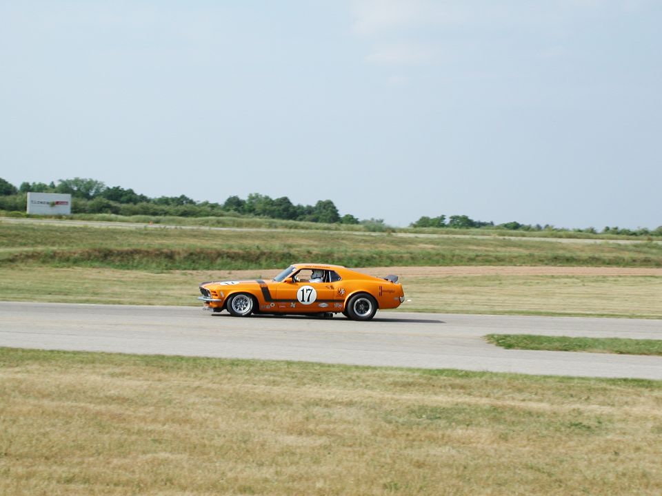 Orange 1970 Ford Mustang race car with black stripes and number 17 on the door, seen from the side on a racetrack