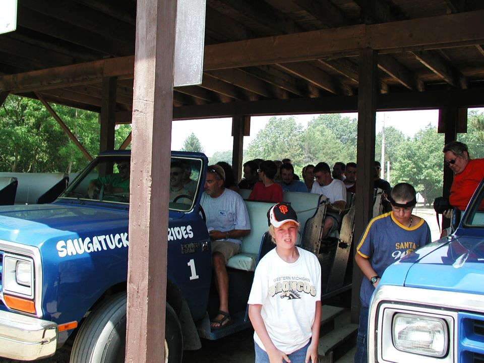 Blue pickup truck with 'SAUGATUCK RIDES' on the side, carrying passengers under a wooden shelter