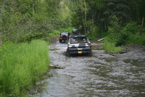 2003 Jeep TJ - Multiple off-road vehicles traverse a wet, muddy path surrounded by trees.