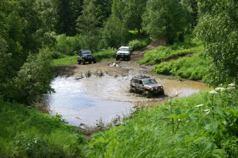 2003 Jeep TJ - Several off-road vehicles navigate a challenging muddy terrain with lush green vegetation surrounding the path.