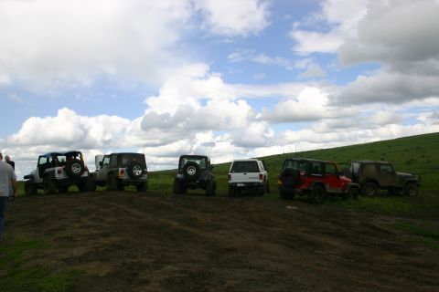 2003 Jeep TJ - A line of Jeeps are parked on a muddy incline with a grassy hill and cloudy sky in the background.