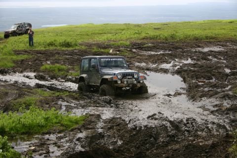 2003 Jeep TJ - A Jeep Wrangler navigates a challenging muddy terrain, with another off-road vehicle and a bystander visible on the higher ground.