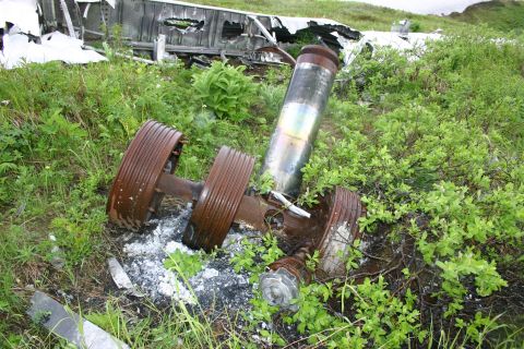 2003 Jeep TJ - Debris from a crashed aircraft is scattered across a grassy area, with a prominent cylindrical component and wheel assembly visible.