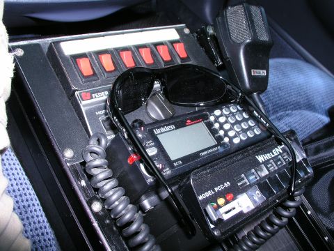 1999 Ford Crown Victoria - Close-up of police radio equipment and siren controls, with sunglasses resting on top.