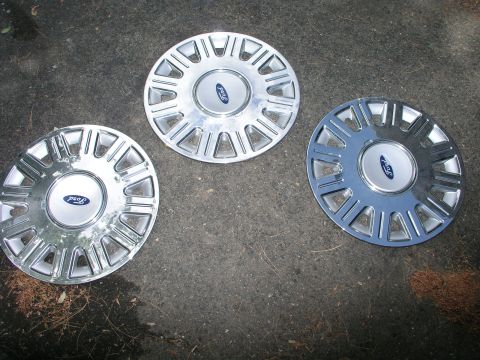 1999 Ford Crown Victoria - Three Ford wheel covers are displayed on a dark, rough surface, with two matching and one slightly varied.