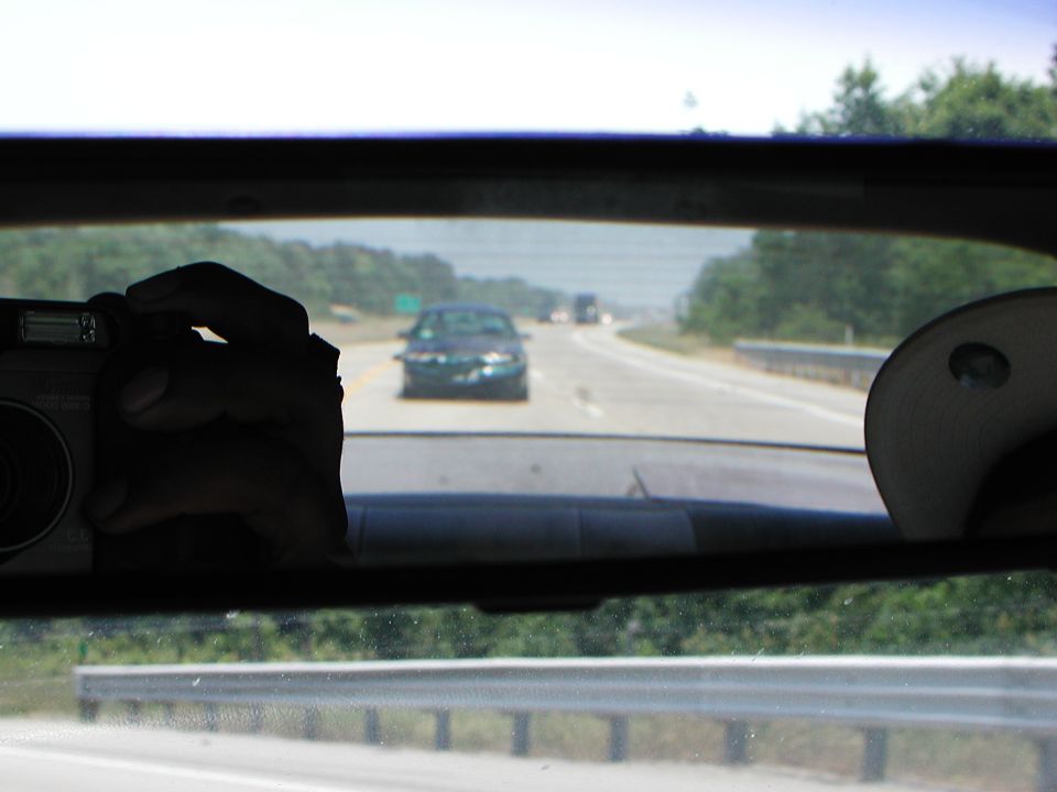 View from inside a car looking through the rear window at a highway with other cars. A hand holds a camera in the foreground