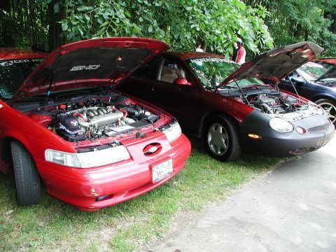 1992 Ford Taurus - A red 1992 Ford Taurus SHO is displayed with its hood open, revealing the V8 engine, alongside other vehicles.