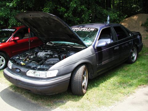 1992 Ford Taurus - A 1992 Ford Taurus sedan is shown with its hood open, revealing the engine compartment. Another red car is visible in the background.