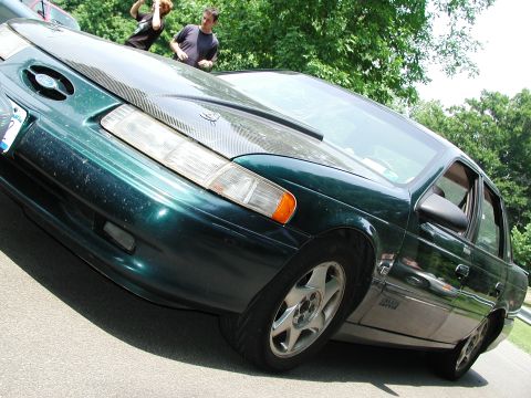 1992 Ford Taurus - A green 1992 Ford Taurus sedan is shown from a low angle, a carbon fiber hood and aftermarket wheels.