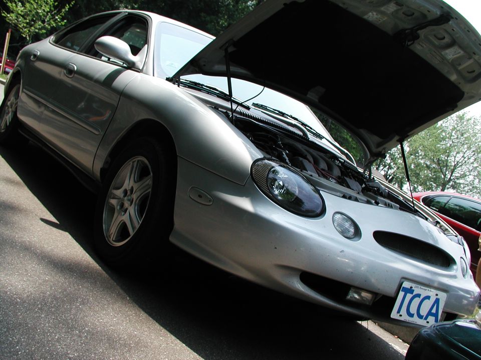 Silver sedan, possibly a Ford Taurus, with hood open showing engine. Another red car is visible behind it