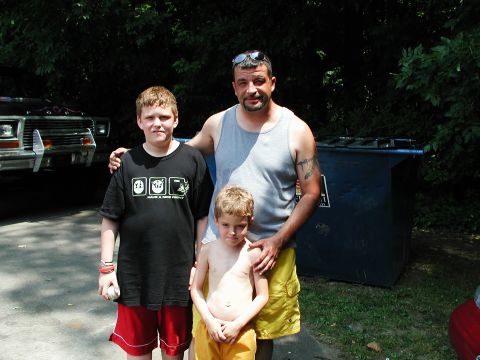 1992 Ford Taurus - A man and two boys stand together outdoors, with a dumpster and part of a vehicle visible in the background.