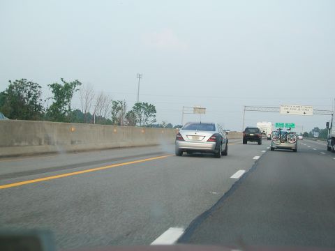 1992 Ford Taurus - A silver sedan is seen on the highway, with smoke coming from its rear. Another vehicle with bicycles is ahead.