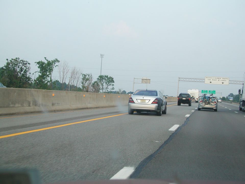 Silver sedan driving on highway, emitting smoke from rear. Another car with bikes on back is ahead
