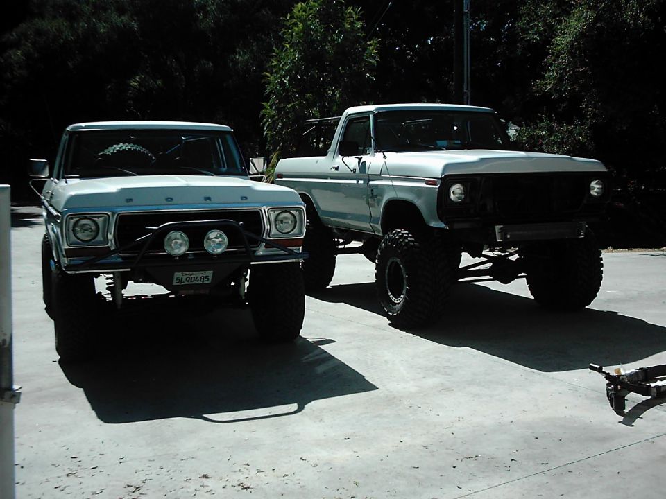 Two lifted Ford trucks parked side-by-side, one light blue with a black grille guard and two fog lights, the other silver with a black grille