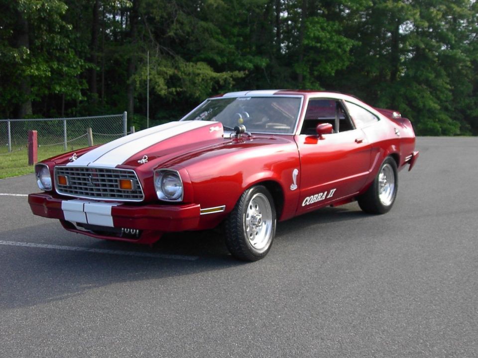Red 1976 Ford Mustang II Cobra II with white racing stripes on hood and front bumper, chrome wheels, and a hood scoop