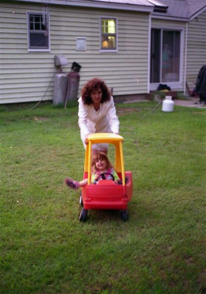 1991 Ford Bronco - A woman pushes a young girl in a red toy car across a grassy yard.