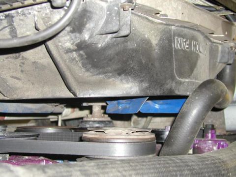1988 Ford Bronco - Low-angle view up into the front of the engine showing the dusty black plastic fan shroud embossed "CAUTION FAN", with the serpentine belt,…
