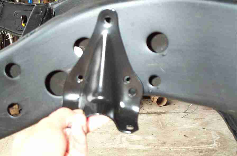 Hand holding a black-painted metal bracket in front of a restored black frame rail in a garage