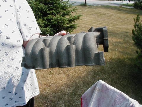 1997 Ford Taurus - Gray metal intake manifold held by a person in front of a grassy yard and trees.
