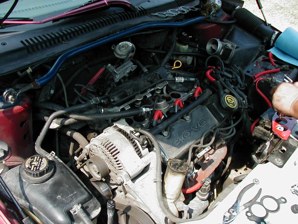 Engine bay of a red car with a 3.0L V6 engine, showing the alternator, battery cables, and various hoses