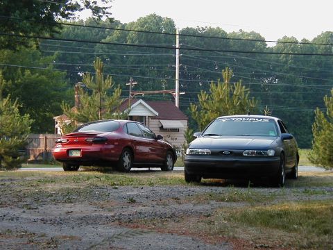 1997 Ford Taurus - Two Ford Taurus sedans are parked in a grassy area with trees and a house in the background.