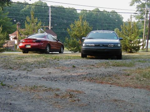 1997 Ford Taurus - Two Ford Taurus sedans are parked in a lot, one black and one red, with trees and houses in the background.