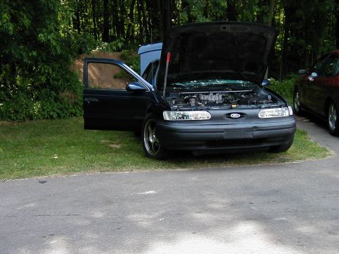 1997 Ford Taurus - A dark blue 1997 Ford Taurus is parked on grass with its hood and driver's door open, with another vehicle visible to the right.