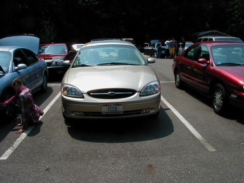 1997 Ford Taurus - A gold Ford Taurus is parked in a lot with other vehicles, one of which has its hood open.