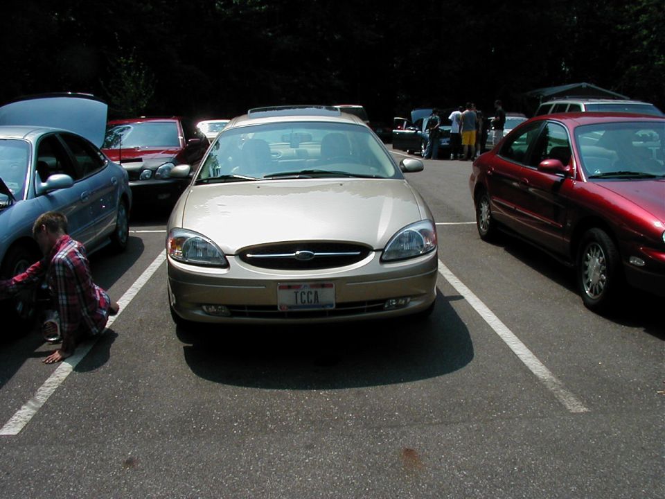 Gold Ford Taurus parked, license plate reads TCCA. Another car is parked to its left with hood up, and a red car is to its right