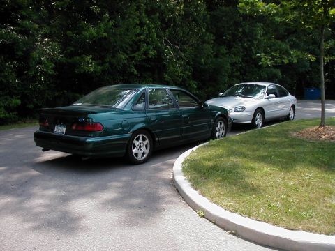 1997 Ford Taurus - Two sedans are parked on a paved area next to a grassy verge with trees in the background.