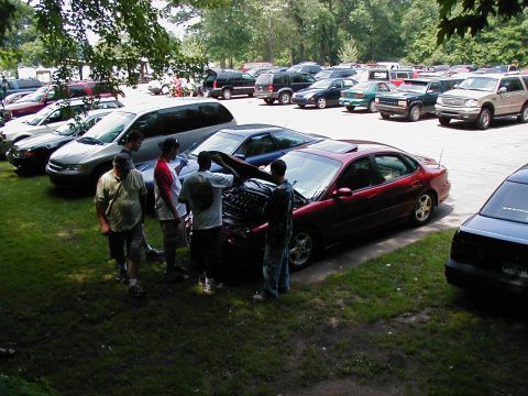 1997 Ford Taurus - A group of people inspect the engine of a red sedan parked in a lot filled with other vehicles.