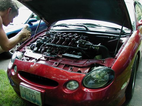 1997 Ford Taurus - The engine bay of a red sedan is open, with a person attending to it. A second vehicle is parked behind.