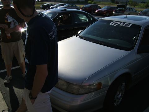 1998 Ford Taurus - A silver sedan is parked in a lot with other cars and people nearby.
